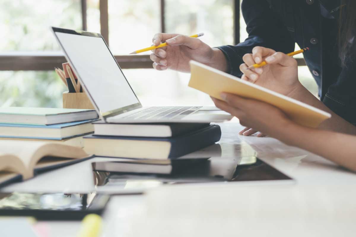 Dos personas al frente de un computador portatil, tomando nota en una libreta y analizando información