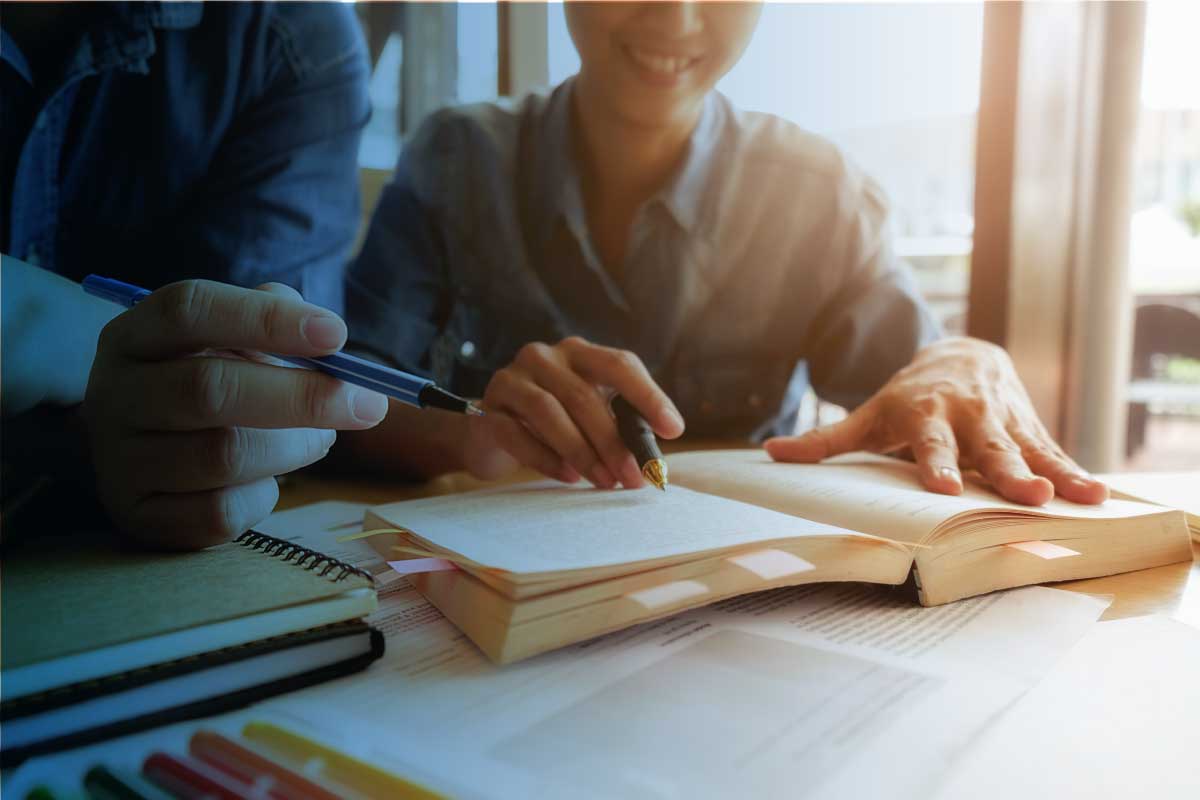 Un hombre y una mujer sentados al frente de un escritorio analizando información de un libro y tomando notas.