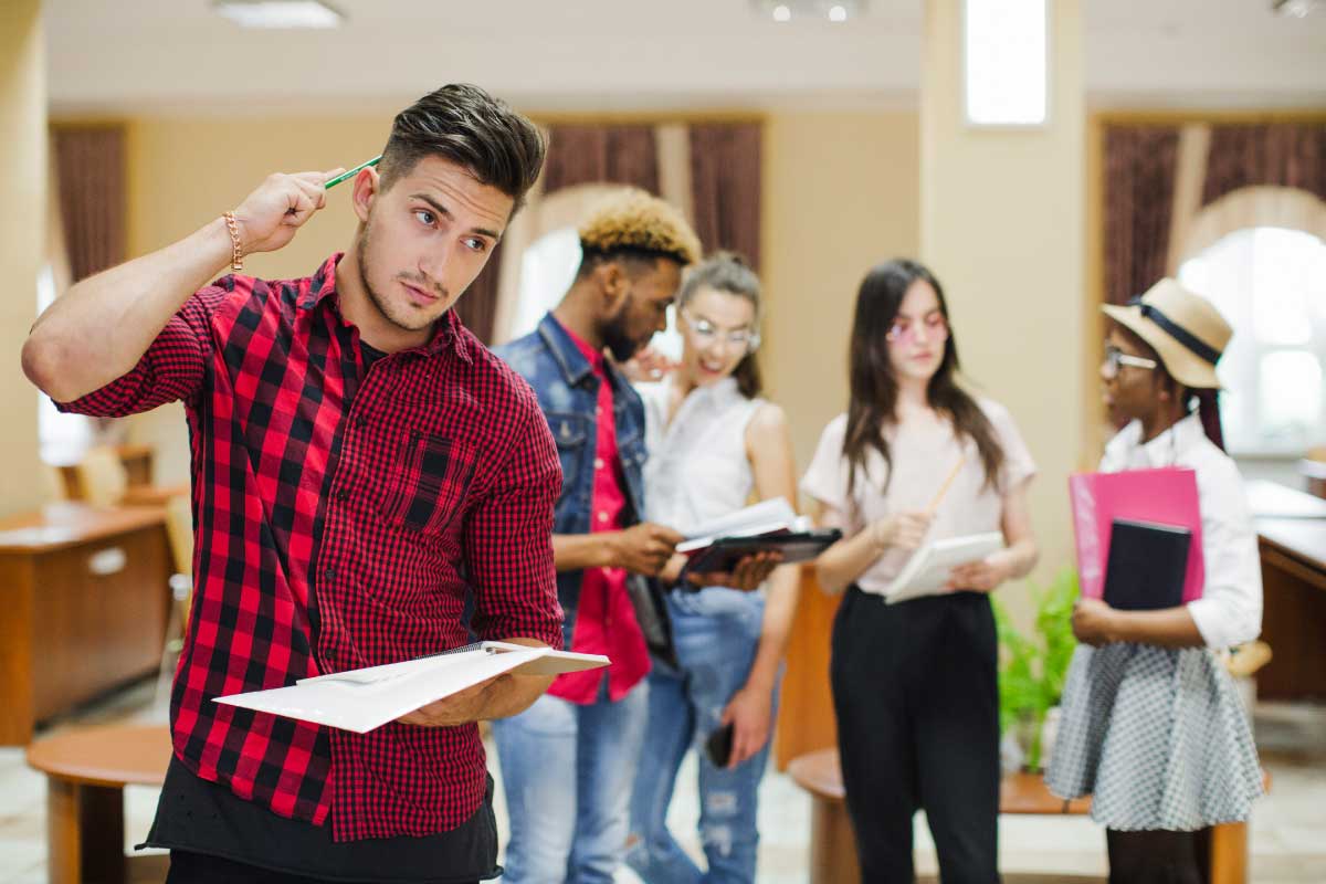 Joven estudiante sosteniendo un cuaderno y un lápiz, al frente de un grupo de estudiantes jóvenes los cuales conversan..