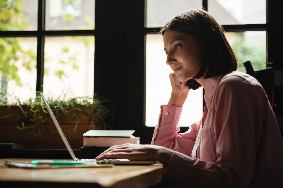 Mujer joven, sentada al frente de un computador portable, con una de las manos sobre el teclado y la otra en su mejilla, pensando.