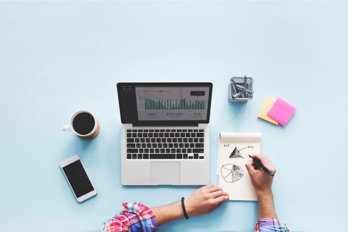 Toma superior de una mesa en la que se ven las manos de una persona haciendo bosquejos gráficas estadísticas, un computador portatil, una taza de café, un teléfono móvil y varios elementos de oficina.n