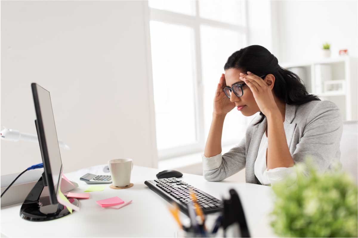 Mujer sentada al frente de un computador de escritorio, con las manos sobre los lados de su frente, mirando preocupadamente.
