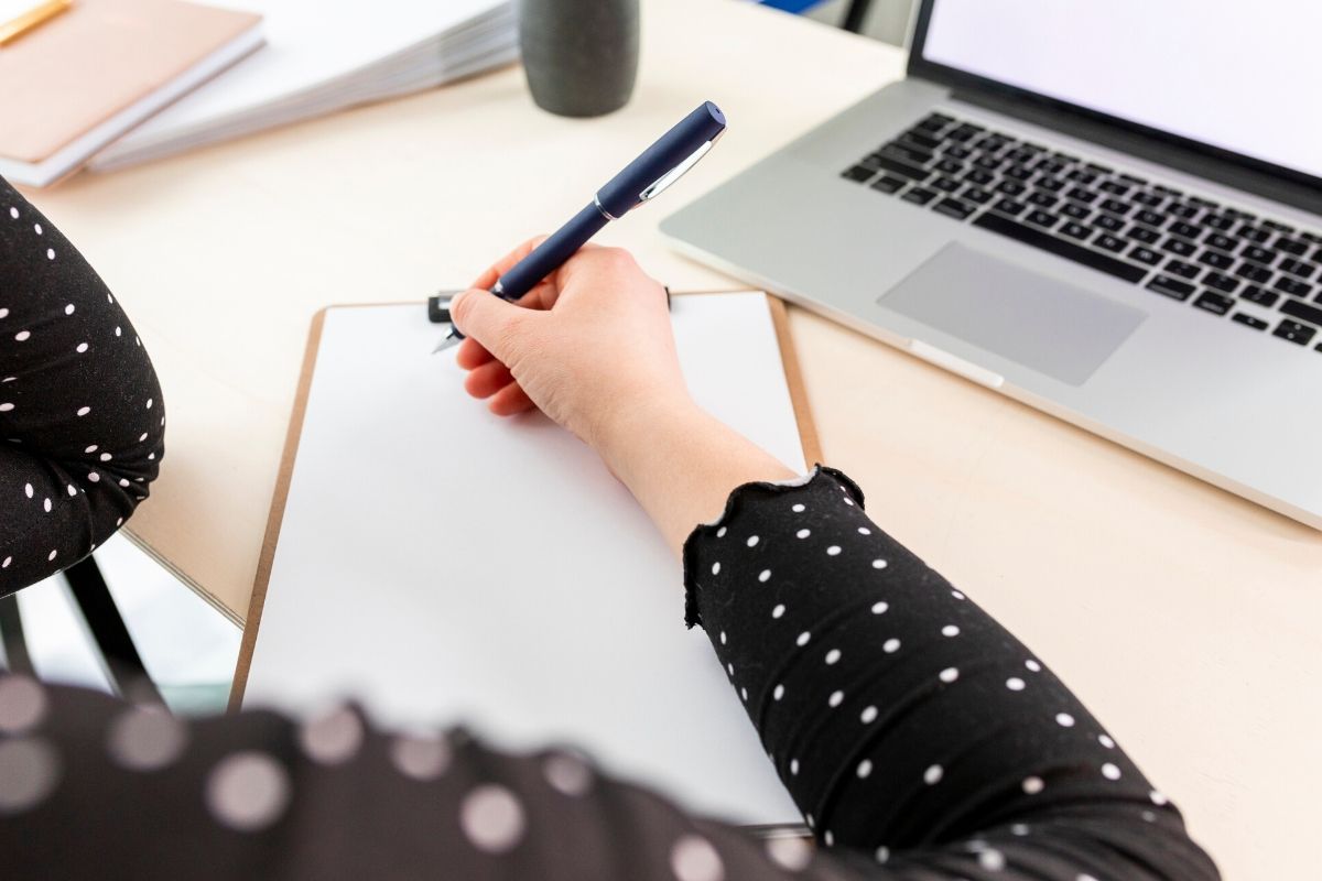Mujer en una mensa escribiendo en una hoja blanca y cerca su computador