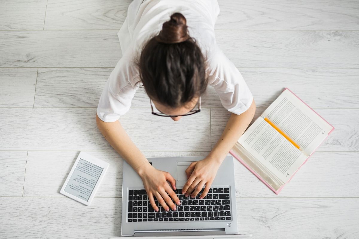 Toma aérea de mujer acostada en el piso boca abajo escribiendo en un portatil y con libros a su alrededor