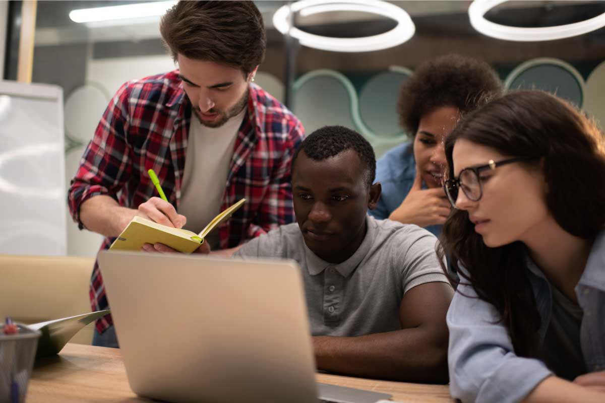 4 compañeros de estudio, sentados alrededor de un computador portatil, uno de ellos tomando nota.