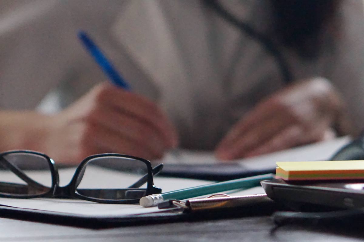 Primer plano de unas gafas y materiales de oficina, con una persona escribiendo de fondo.
