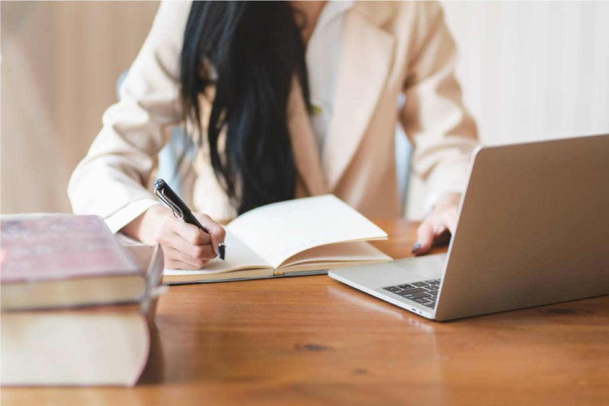 Fotografía de una mujer a la cual no se le ve la cara, escribiendo en una cuaderno con un computador al frente.