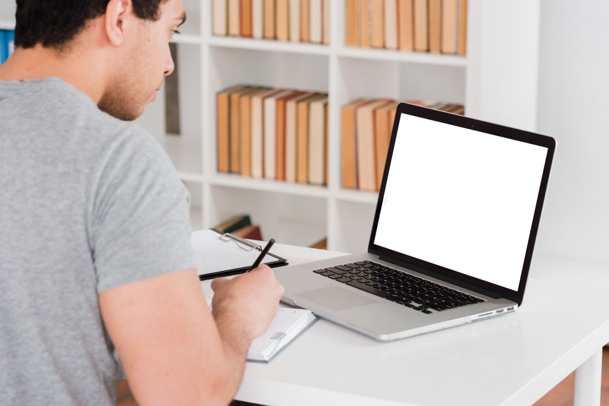 Hombre joven sentado al frente de un computador portatil, mirando la pantalla y tomando notas en una libreta.