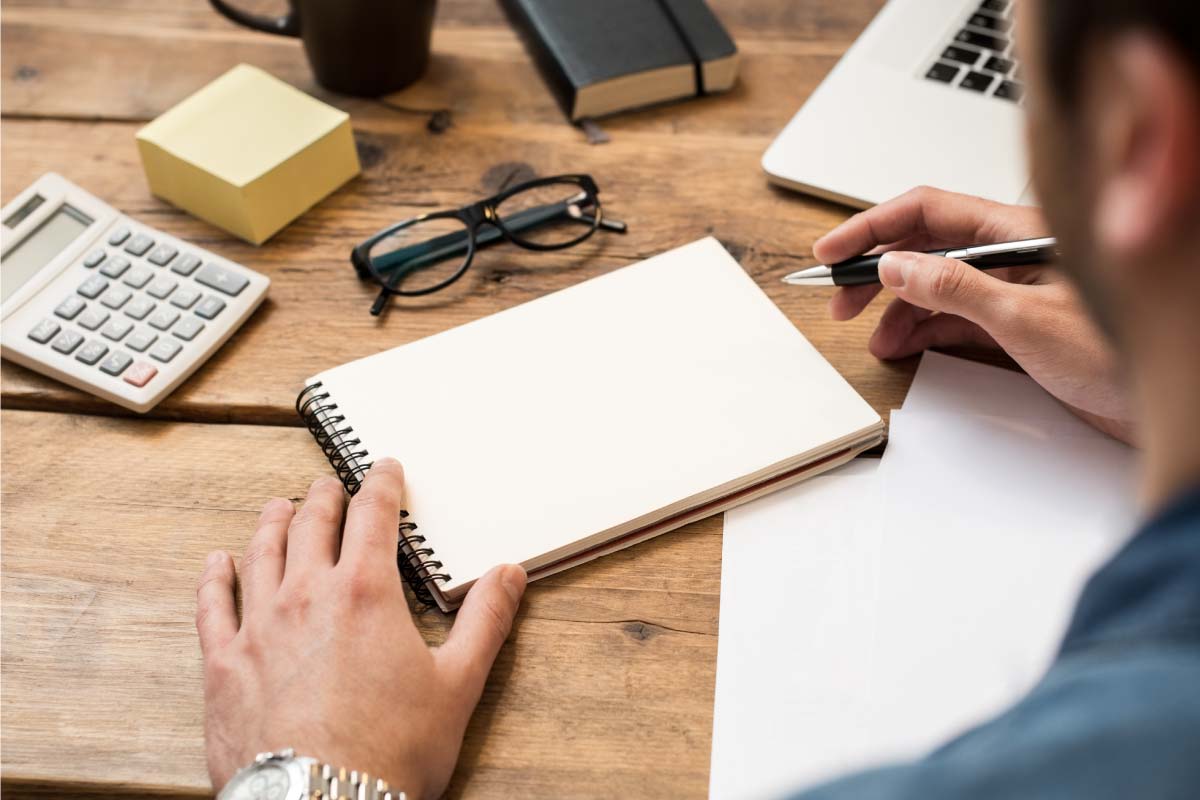 Toma trasera de un hombre sentado al frente de un escritorio, escribiendo en una libreta.