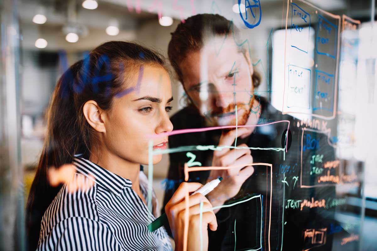 Un hombre y una mujer escribiendo sobre un tablero transparente desarrollando una idea.
