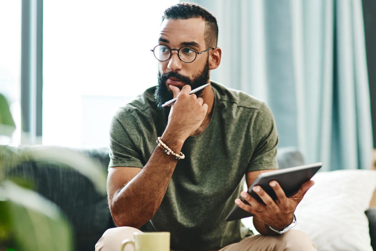 Hombre joven, sentado en un sofá y sosteniendo una tableta, mirando a lo lejos con su mano en el mentón.