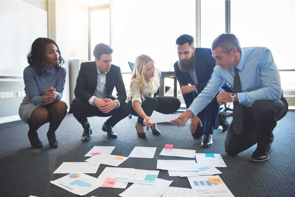 Grupo de cinco empleados de oficina analizando papeles financieros en el suelo