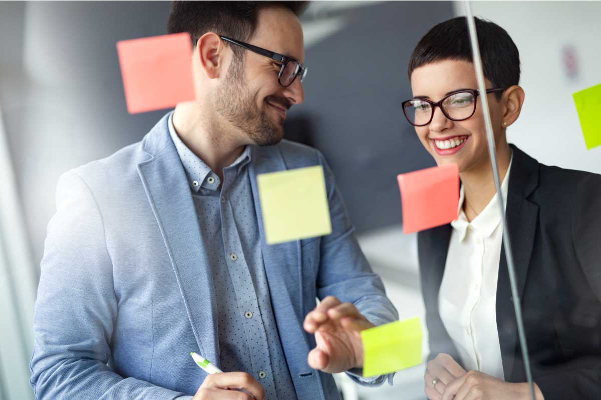 2 profesionales jóvenes sonriendo, al frente de un tablero transparente con post its en él.
