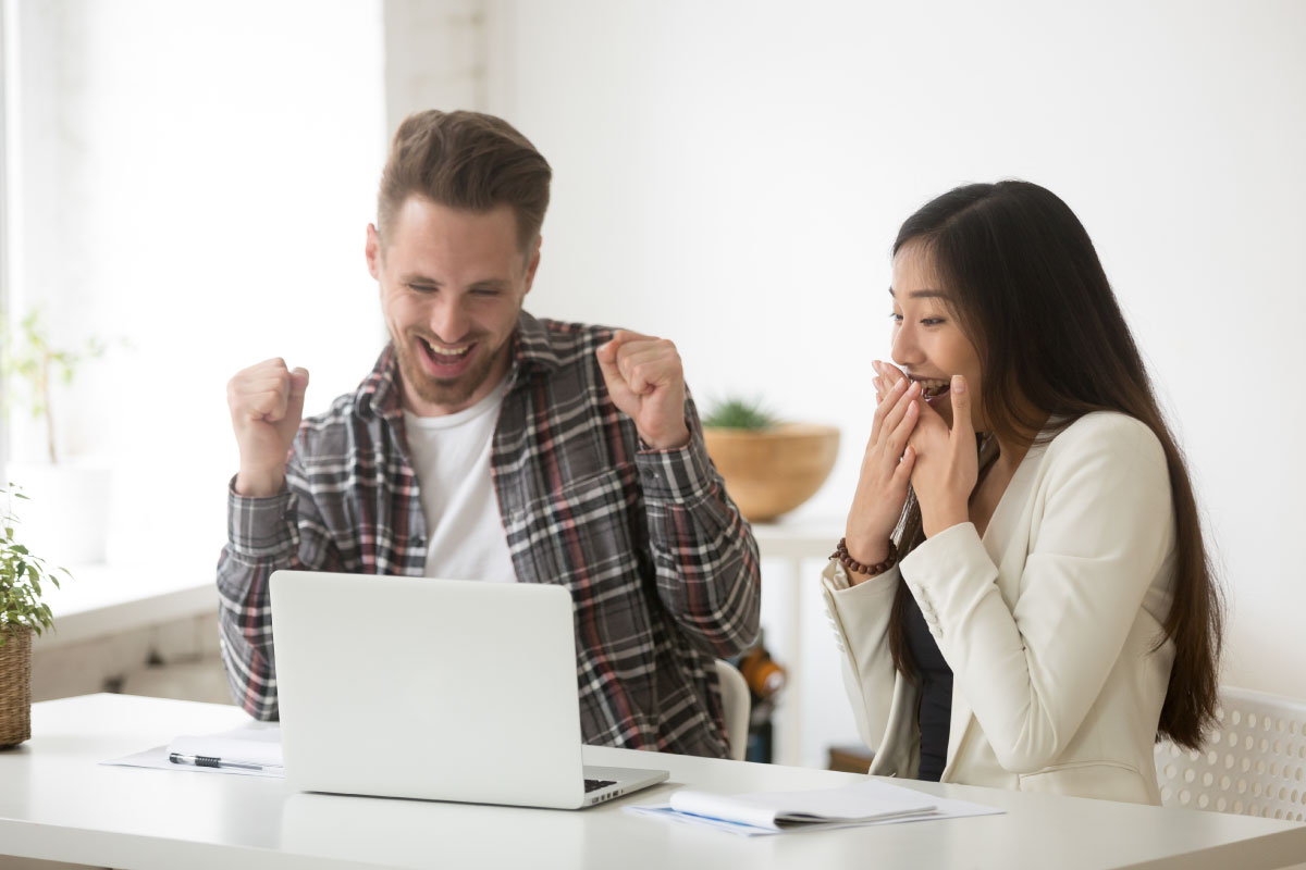 Un hombre y una mujer jóvenes, sentados al frente de un computador portatil, sonriendo y celebrando.
