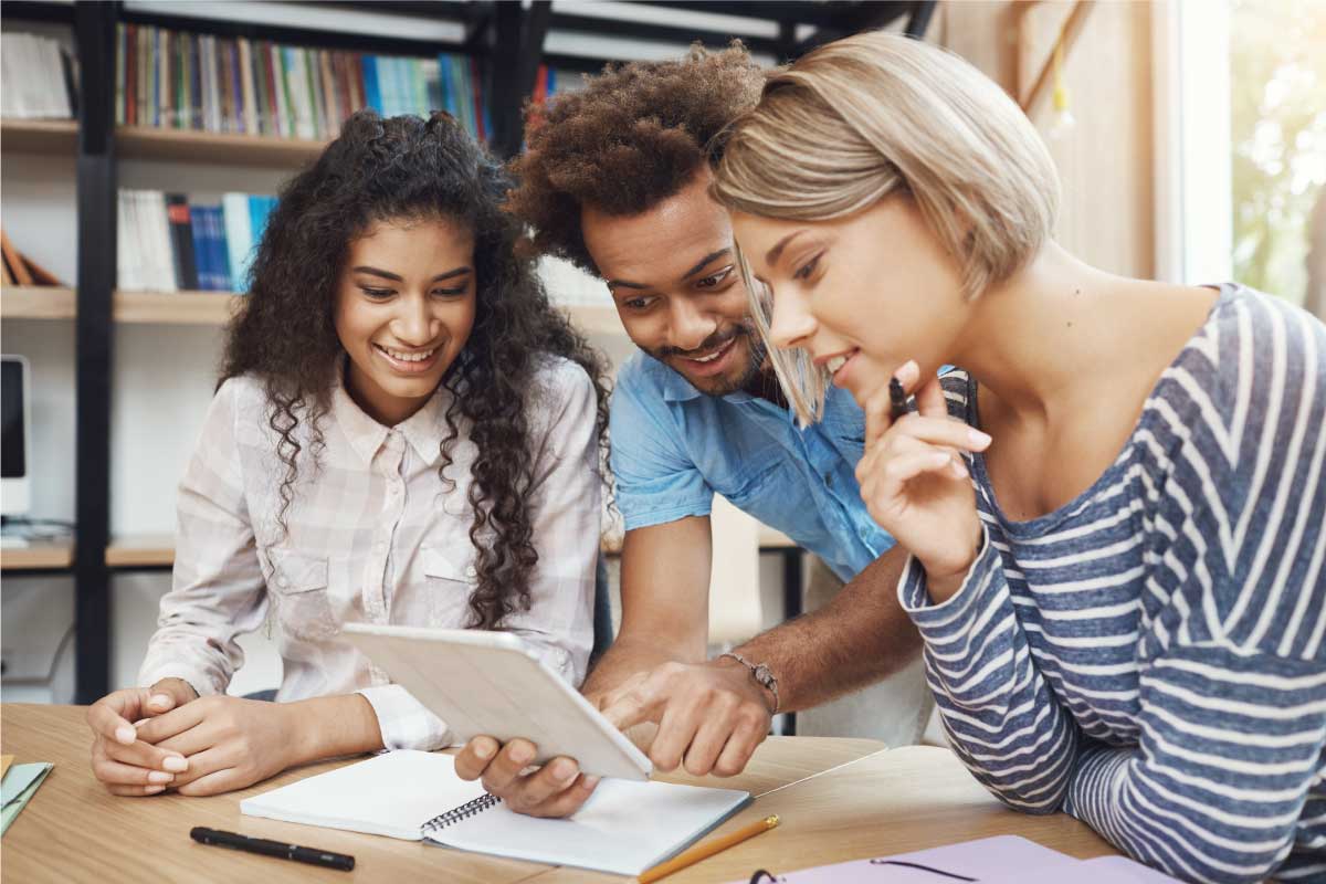 Grupo de tres personas jóvenes, sentadas, mirando una tableta que la persona del centro sostiene.