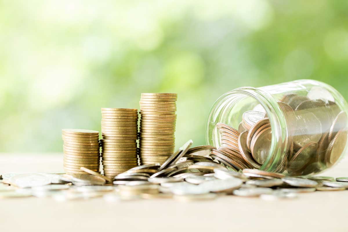 Acercamiento de un conjunto de monedas esparcidas sobre una superficie, un grupo de ellas formando 3 pilas, y un recipiente de vidrio, volteado con monedas adentro