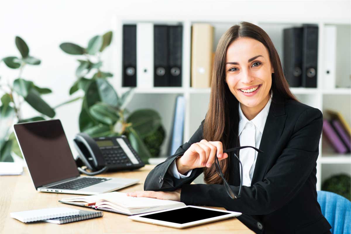 Mujer de negocios sentada en un escritorio, mirando hacia la cámara y sosteniendo sus lentes.