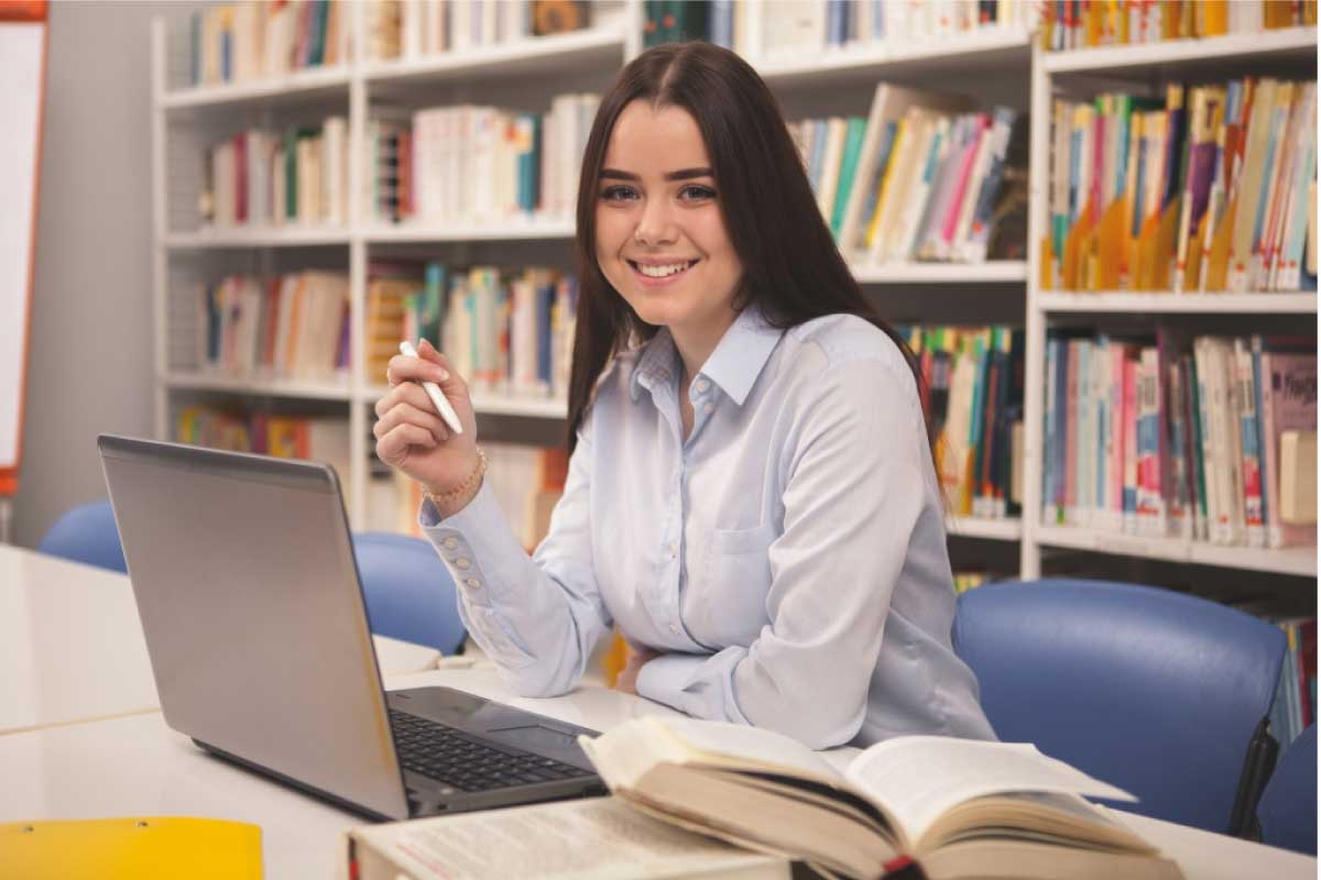 Mujer sonriendo, sentada al frente de un computador portatil en una biblioteca.
