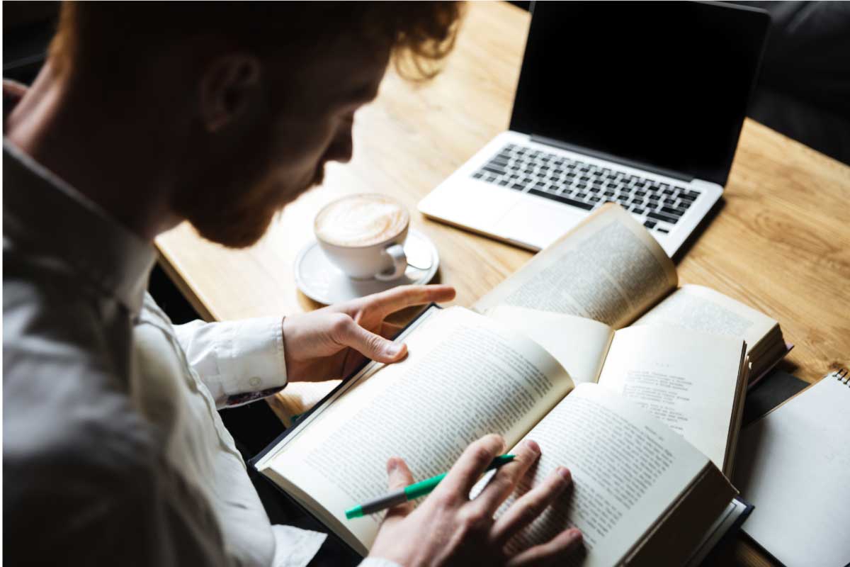 Hombre joven sentado al frente de un computador portatil, con un libro abierto y un lápiz en su mano, dispuesto a tomar notas.
