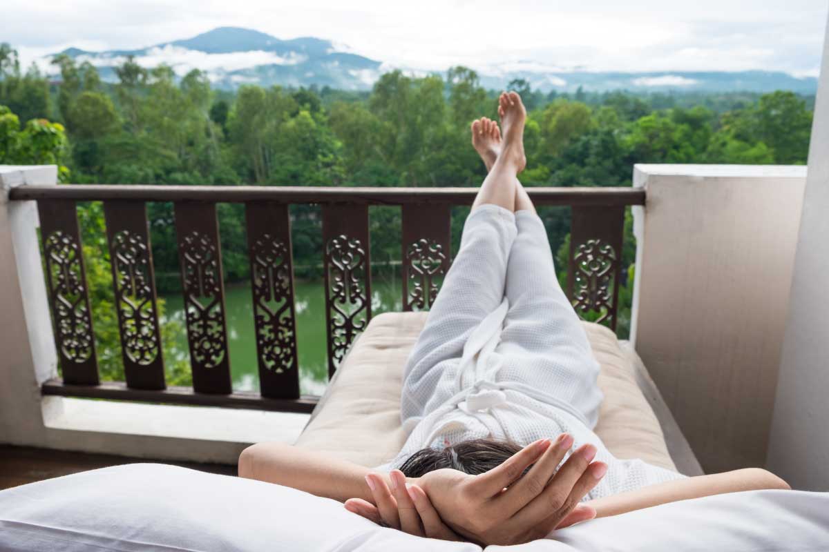Mujer acostada en un sofa en una terraza, con montañas y bosques en la distancia.
