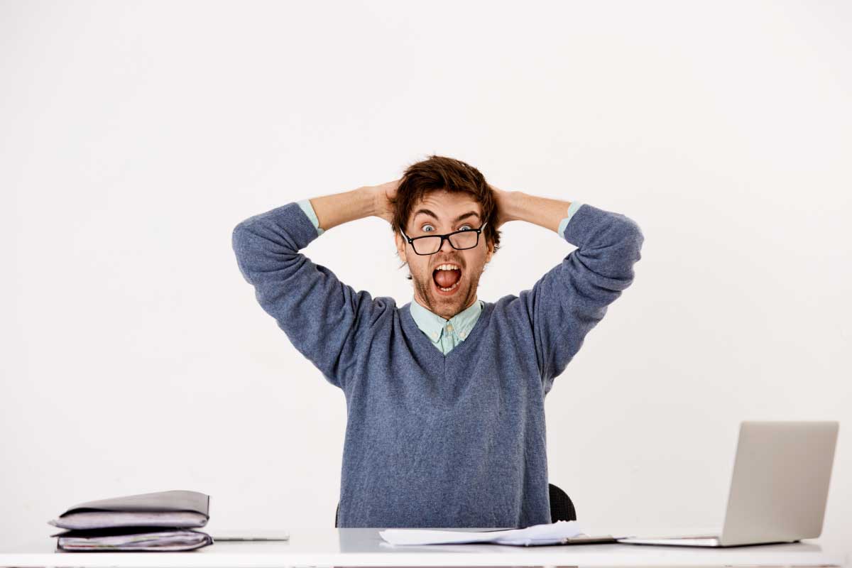 Hombre con gafas torcidas, boca abierta y manos detrás de la cabeza, sentado al frente de un computador
