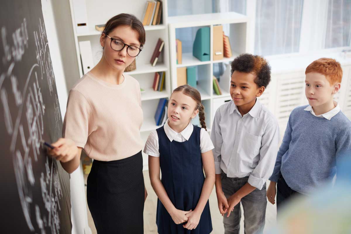 Profesora y tres alumnos parados al frente de un tablero negro, explicando un concepto.