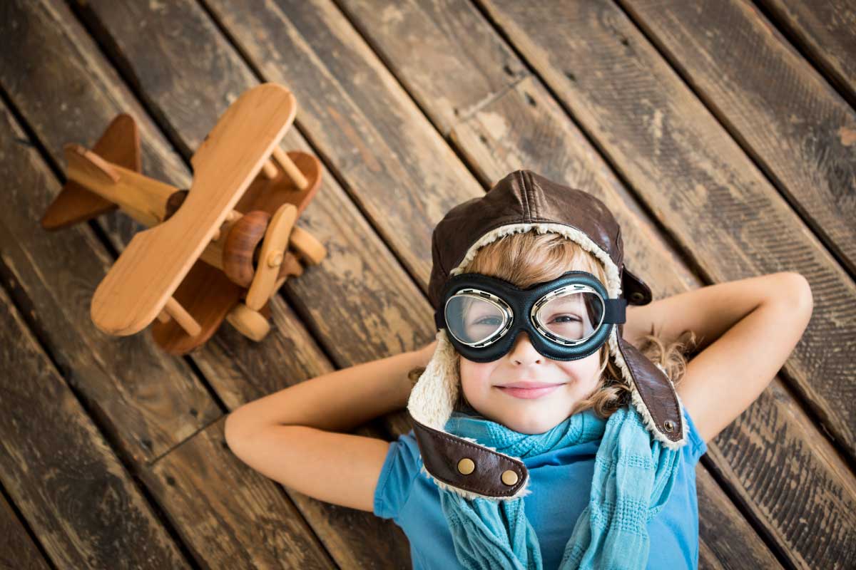 Niño vestido con gorra y gafas de piloto, acostado sobre un piso de madera con sus manos bajo su cabeza y sonriendo.