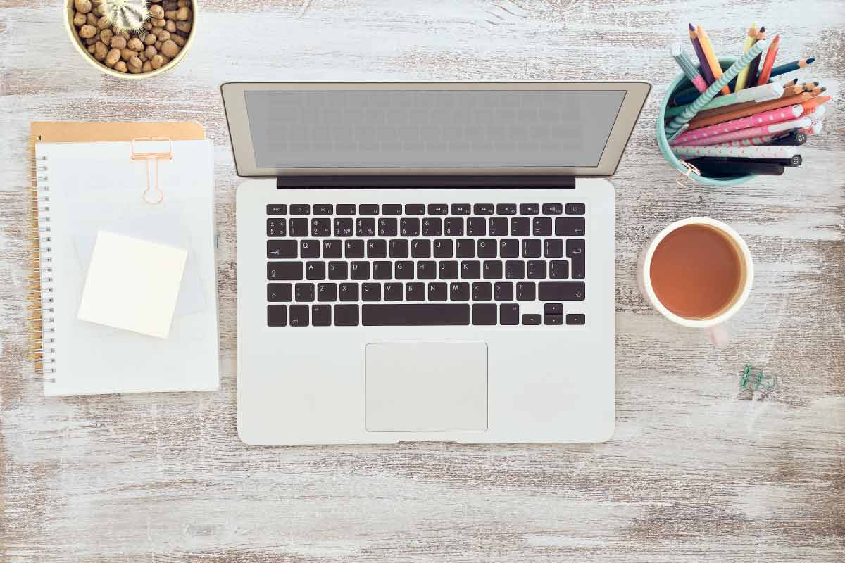 Vista aérea de un escritorio en el que se encuentra un computador portable abierto, una libreta, una bebida y unos lápices de colores dentro de un recipiente.