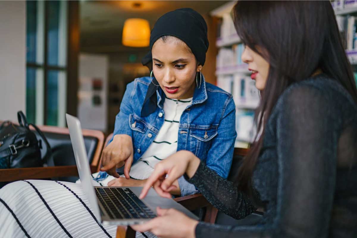 Dos mujeres jóvenes, sentadas al frente de un computador portatil, señalando la pantalla.