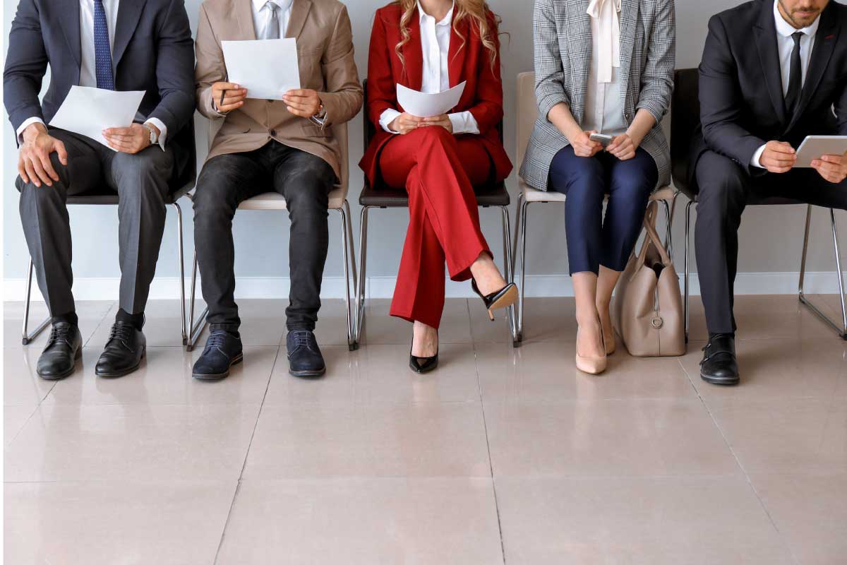 Vista frontal de 6 personas, a las que no se les ve la cabeza, sentadas en una sala de espera y leyendo su hoja de vida.