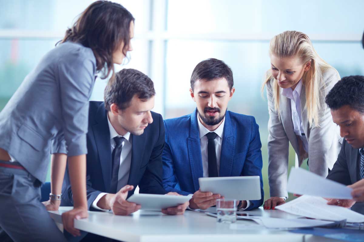 Cinco persona ejecutivas, al rededor de un escritorio mirando una tableta y analizando papeles con información.