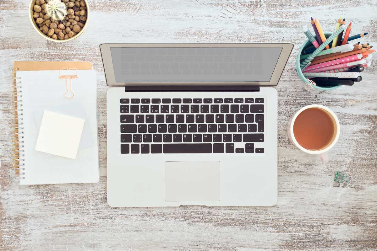 Toma superior en donde se ve un computador portatil, un cuaderno, lápices dentro de un recipiente y un café.