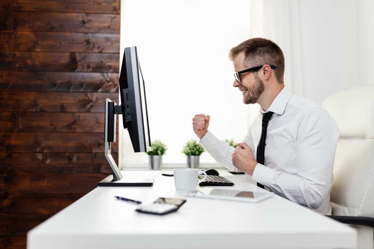 Hombre de negocios sentado al frente de un computador, con sus manos empuñadas celebrando una victoria.