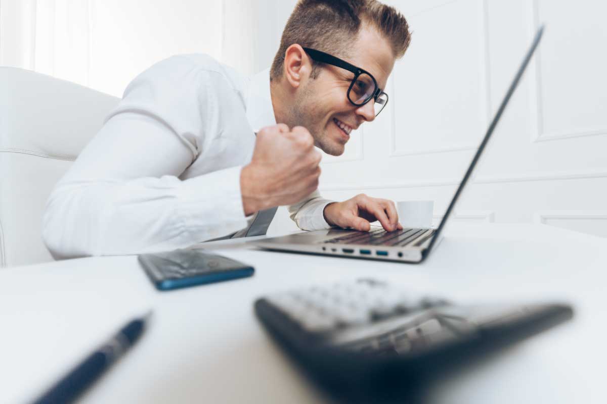 Hombre de negocios sentado al frente de un computador portatil, con el puño cerrado y celebrando.