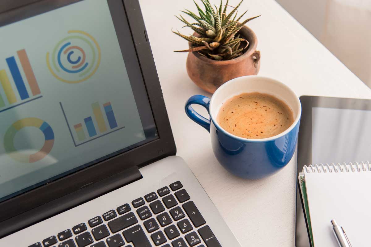 Acercamiento de un escritorio en el cual hay un computador portatil, una taza de café, una planta pequeña y una libreta de notas