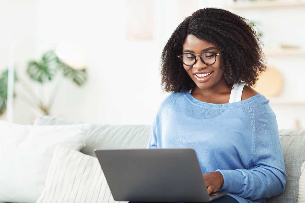 Mujer joven sentada en un sofá con un computador portatil sobre sus piernas, escribiendo en él.