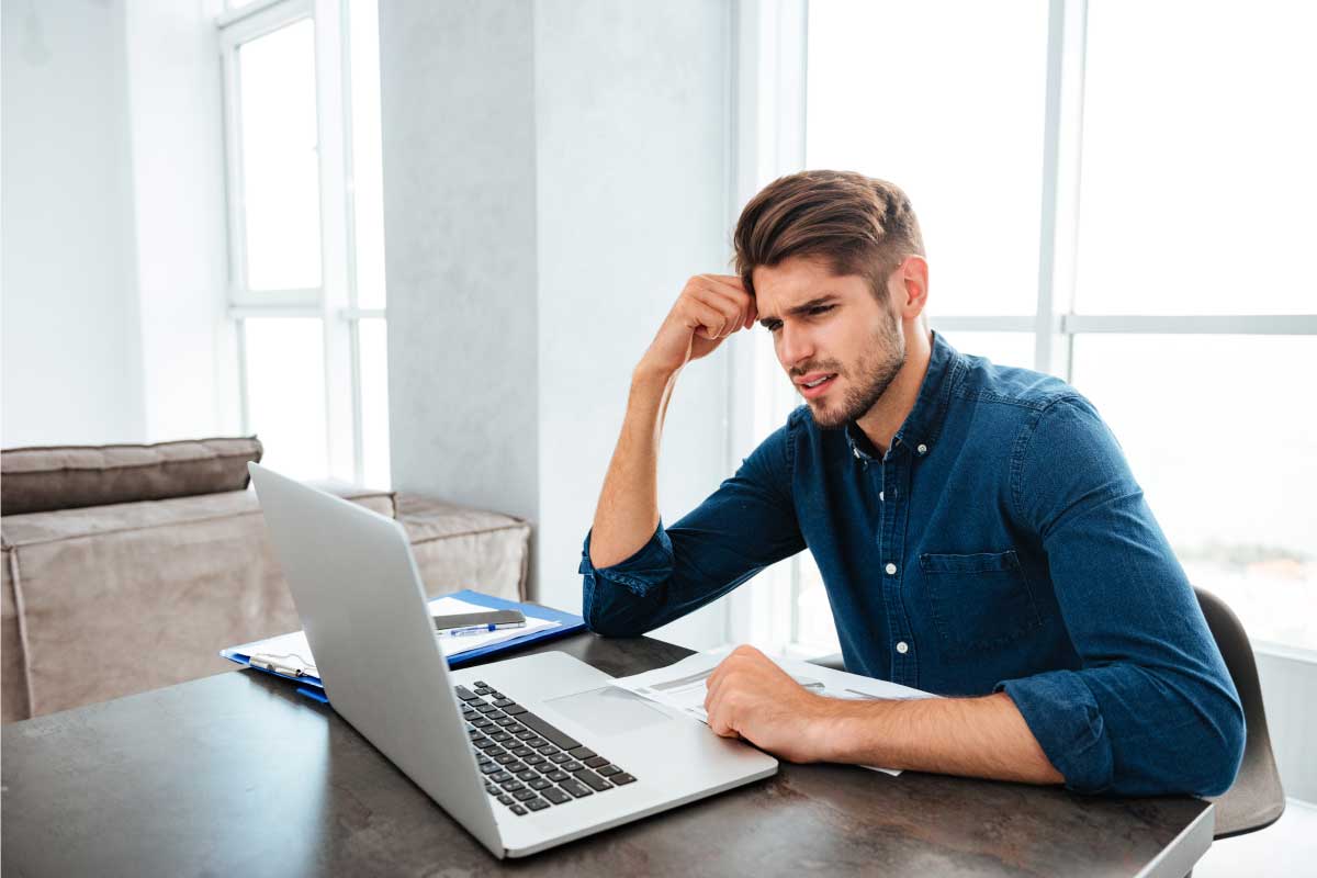 Hombre joven sentado al frente de un computador portatil, con los codos sobre la mesa y el puño derecho en su frente preocupado.