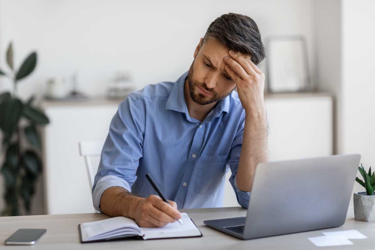 Hombre joven sentado al frente de un escritorio, con un computador portable abierto y tomando nota en una libreta.