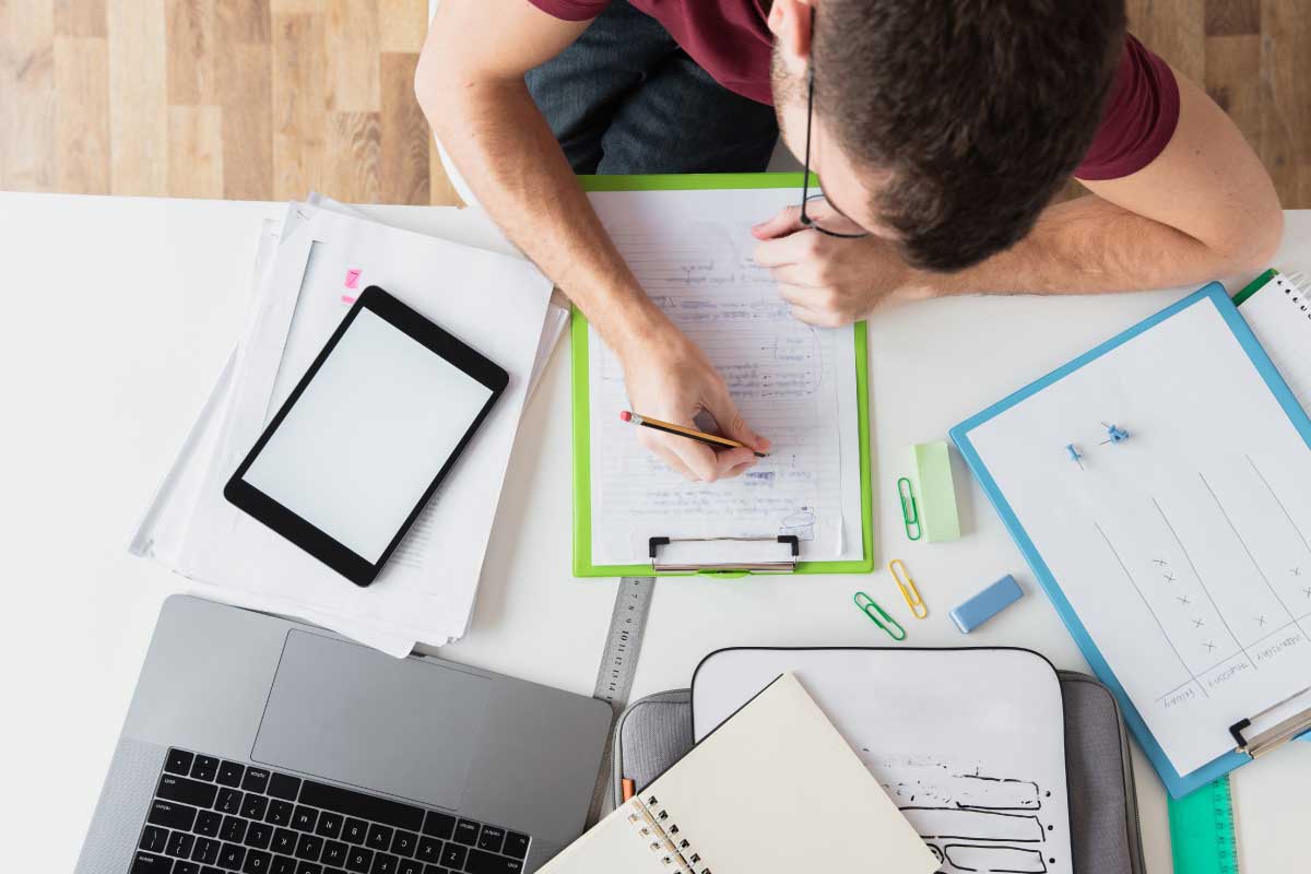 Toma superior de un hombre sentado en un escritorio escribiendo en una libreta donde además se ve un computador y varios papeles