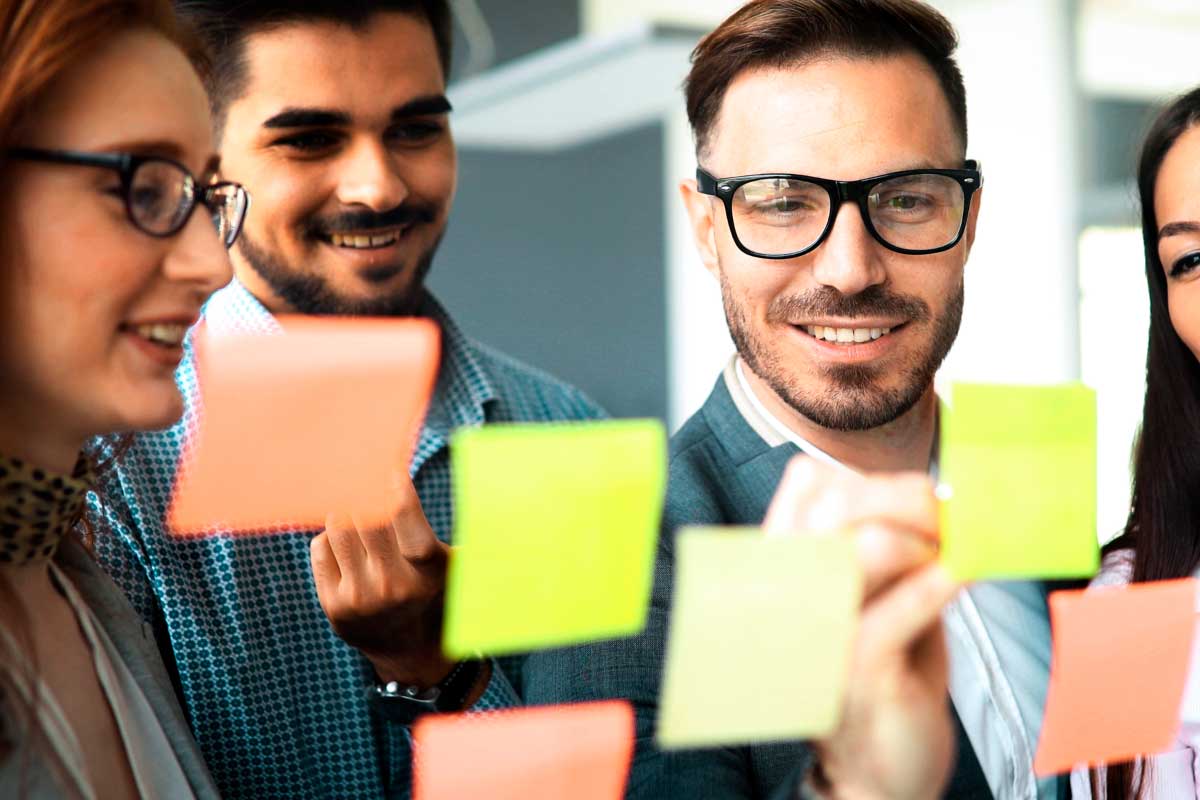 Grupo de jóvenes ejecutivos sonriendo, al frente de un tablero transparente tomando notas sobre post-its