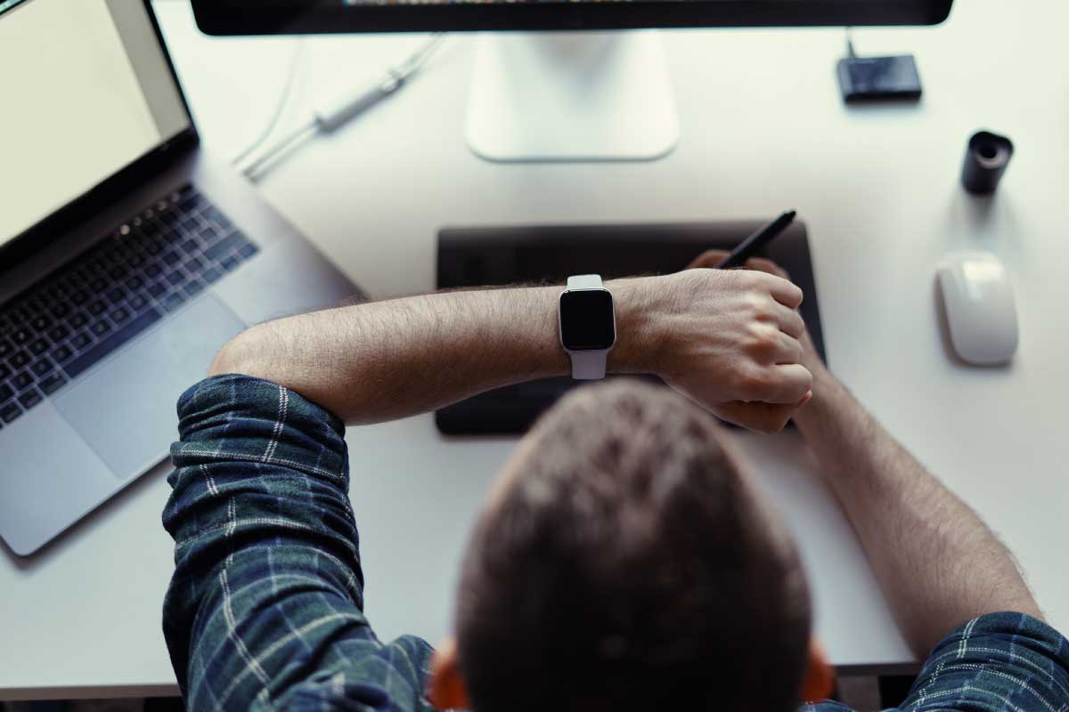 Toma superior de un hombre sentado en un escritorio, con la mano levantada la frente mirando su reloj.