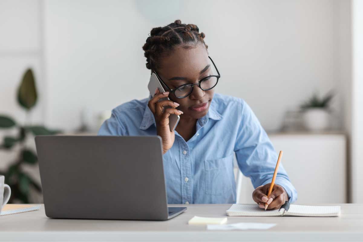 Mujer sentada al frente de un computador portatil, hablando por un teléfono móvil y tomando nota en una libreta.