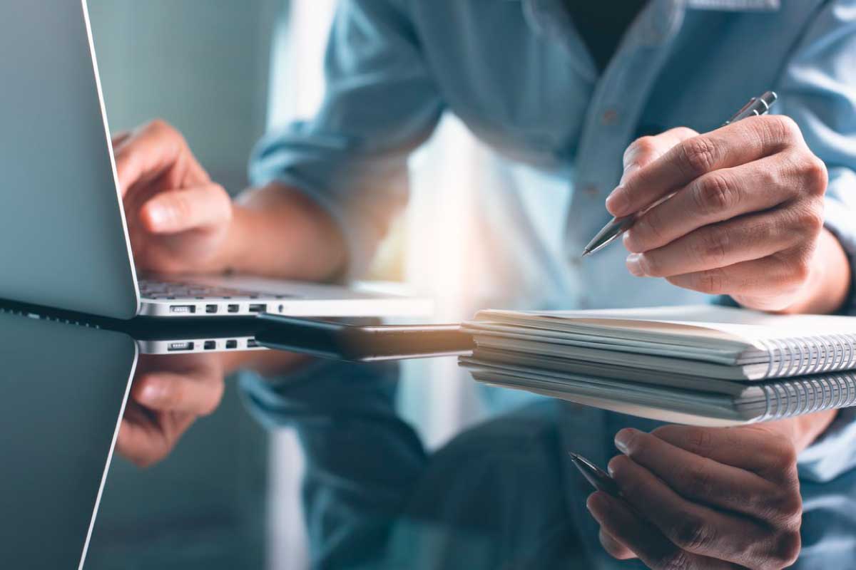 Acercamiento de la manos de una mujer, una mano en un teclado y la otra escribiendo en un cuaderno