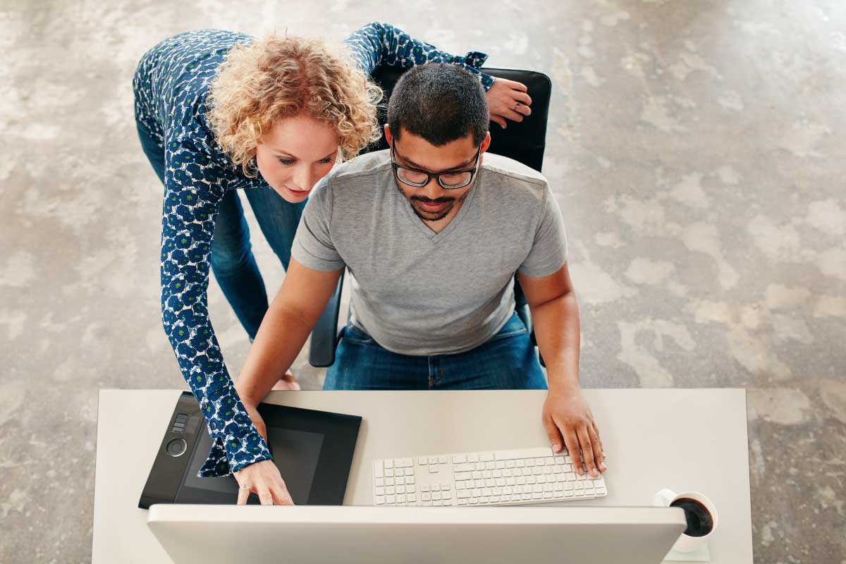 Vista aérea de un hombre sentado al frente de un escritorio y una mujer parada a su lado, analizando información en una pantalla de un computador