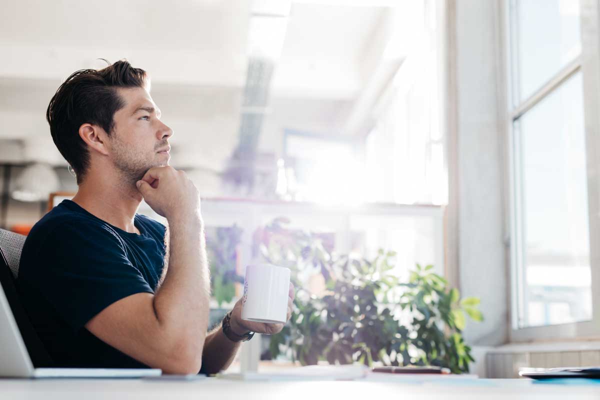 Hombre joven con una mano sobre su mentón y la otra sosteniendo una taza, mirando a través de una ventana.
