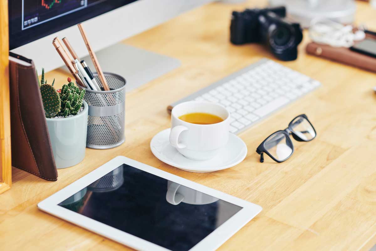Toma lateral de un escritorio sobre el cual hay una tableta, un café, unos anteojos y un monitor y teclado de computador.