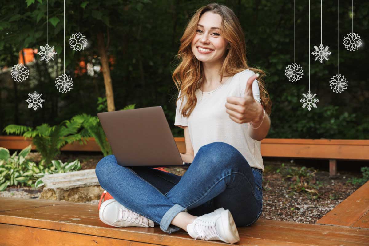 Mujer joven sentada en el piso con los pies cruzados, con un computador portatil sobre las piernas y haciendo la seña de pulgar hacia arriba.