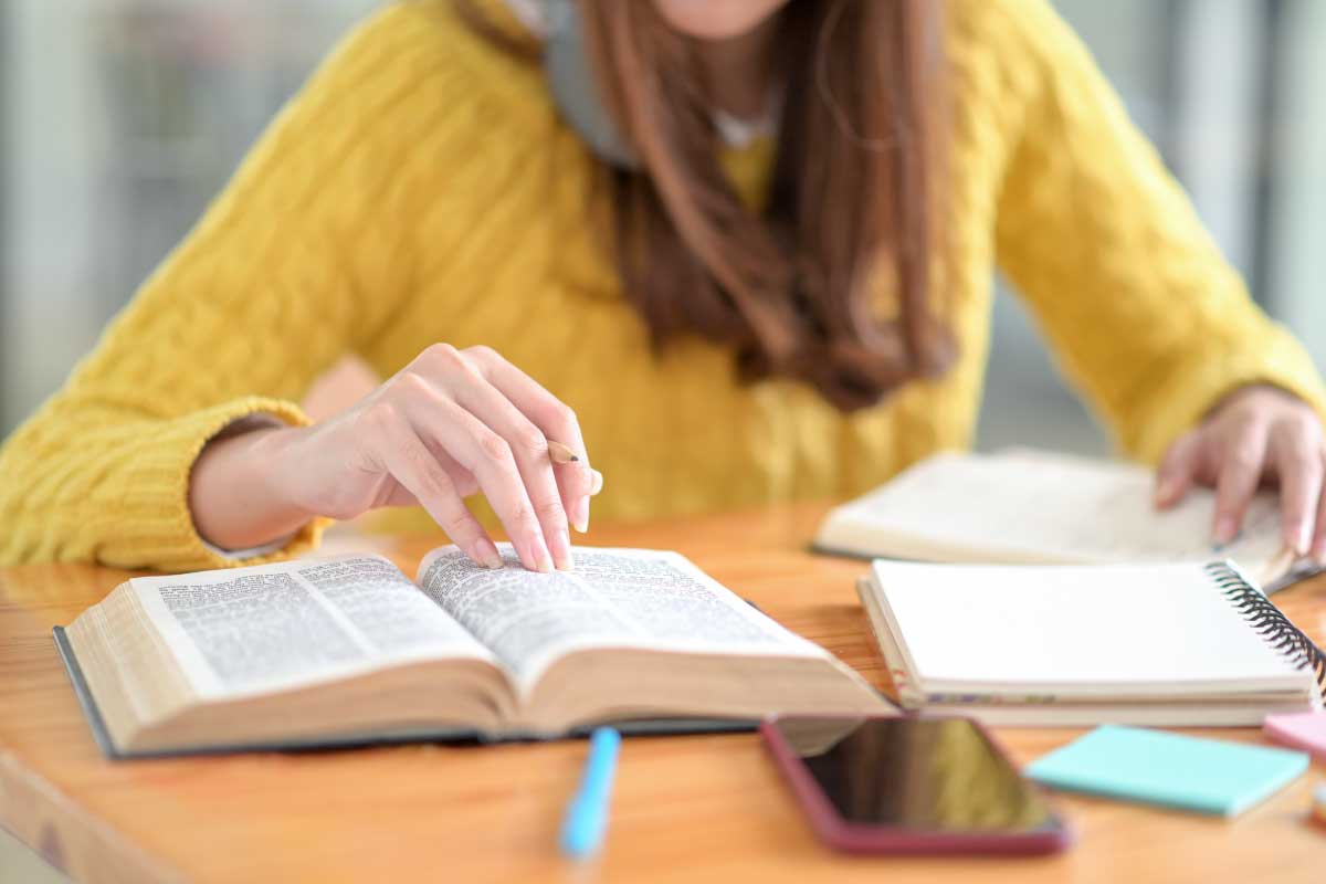 Fotografía de una mujer, donde solo se ve su torso, sentada en un escritorio, revisando un libro y tomando notas.