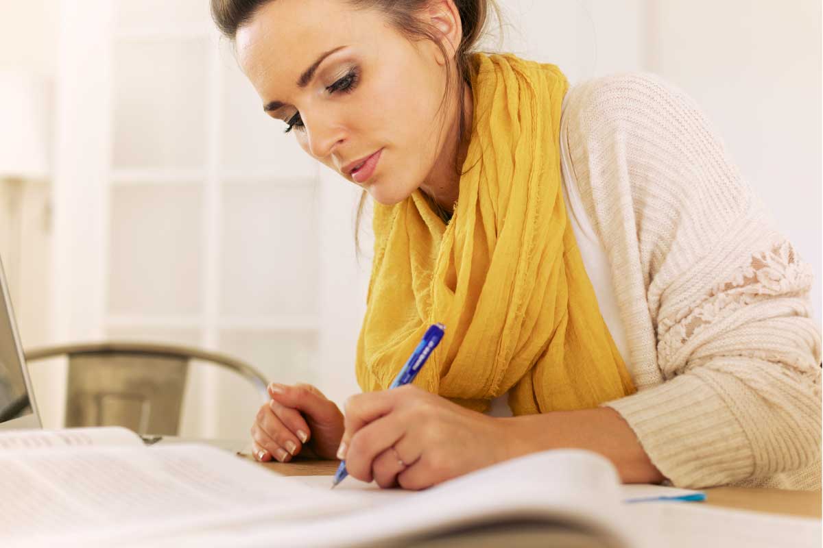 Mujer joven sentada al frente de una mesa estudiando y tomando notas.