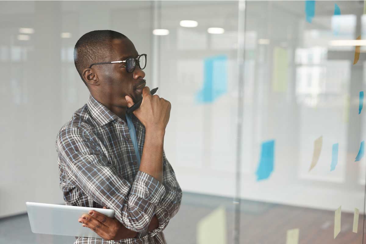 Hombre pensando, con su mano sobre su boca, mirando un tablero con notas.
